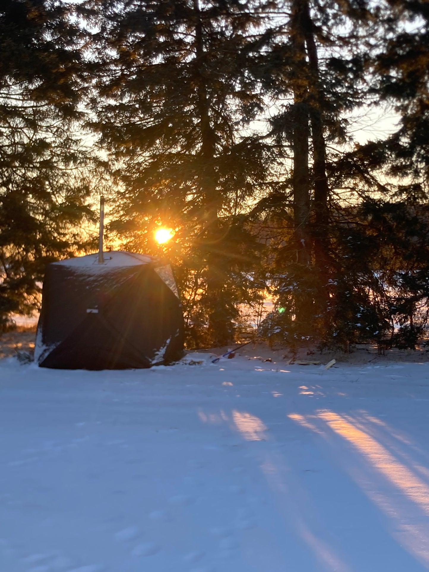 A portable sauna tent with a stove set up in a snowy outdoor environment with sunset in the background.